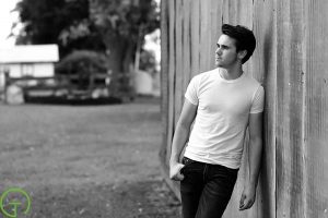 A high school senior boy leans against a rustic barn